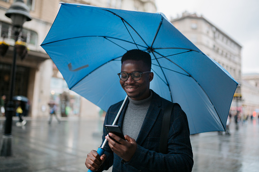 Happy young African American man walking around the city on a rainy day, holding an umbrella, smiling and using a smart phone