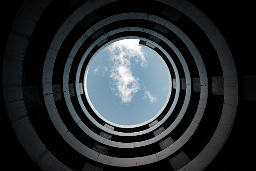 Modern Architecture Spiral Driveway of Urban Parking Garage from below against blue sky with fluffy clouds flying by. Modern Urban Architecture Detail.