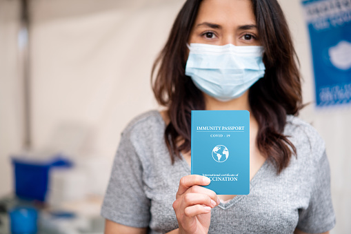 Portrait of a young Latin American woman at a vaccination stand holding a COVID-19 immunity passport certifying her vaccine status. **DESIGN ON PASSPORT WAS MADE FROM SCRATCH BY US**