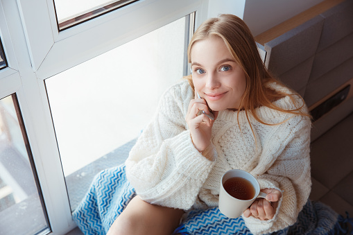 Young blonde woman indoors sitting on window sill wearing woolen sweater holding cup with a hot tea answering phone call smiling