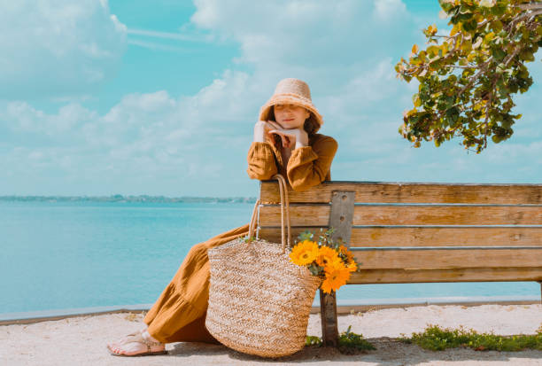 Young woman in beautiful dress sits on bench by the lake and enjoys the view rest on a sunny day. Portrait of a romantic girl in a straw hat against a background of blue water Young woman in beautiful dress sits on bench by the lake and enjoys the view rest on a sunny day. Portrait of a romantic girl in a straw hat against a background of blue water. straw bag stock pictures, royalty-free photos & images