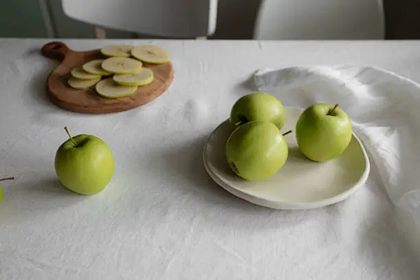 Sliced apples and green apples on a table with a white linen cloth, linen napkin and a handmade ceramic plate, casually arranged to tell the story of preparing and eating a healthy snack during the day.