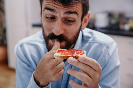 Cute close up of a bearded guy licking his finger while looking at the toast with his favorite jam with joyful smile on his face