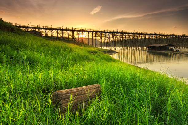 pradera verde junto al río con un puente de madera sobre el río al atardecer en sangklaburi, provincia de kanchanaburi, tailandia - sangkhlaburi fotografías e imágenes de stock