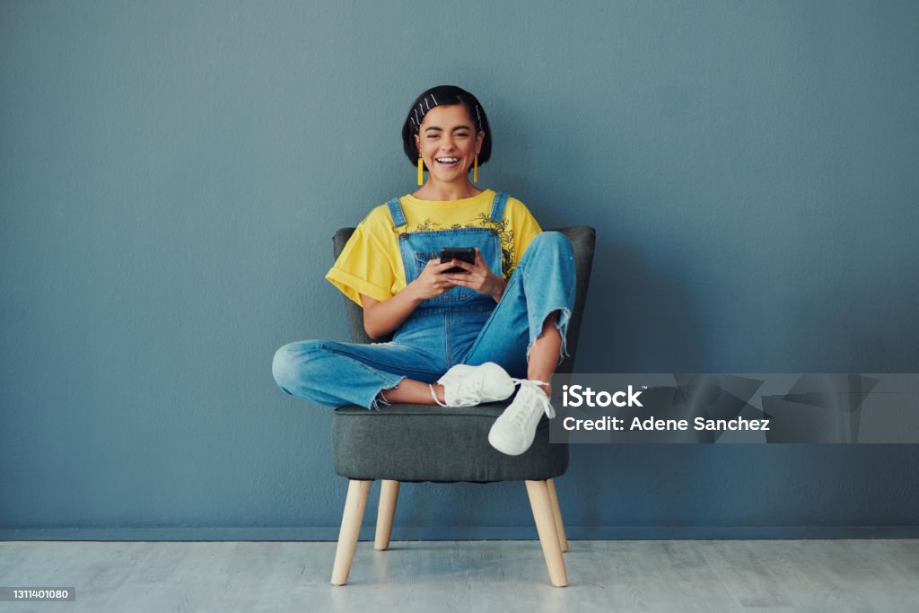 I spread happiness through my memes Shot of a woman using her cellphone while sitting on a chair against a blue wall Teenager Stock Photo