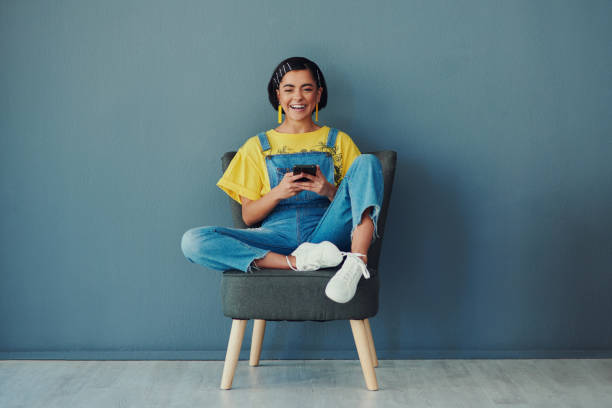 I spread happiness through my memes Shot of a woman using her cellphone while sitting on a chair against a blue wall sitting stock pictures, royalty-free photos & images
