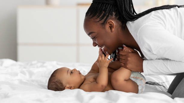 African American mom playing in bed with her black baby Speech And Language Development For Babies. Closeup profile portrait of beautiful little black infant smiling, young black woman playing and holding kid's legs in hands, banner, free copy space african american stock pictures, royalty-free photos & images
