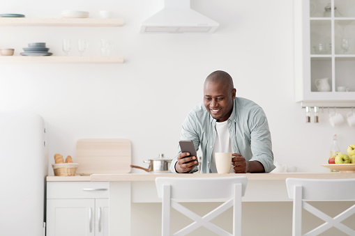 Happy african american man using smartphone and having morning coffee, standing in the kitchen, texting on cellphone, surfing internet and reading news, free space