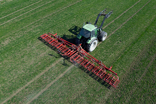 Aerial view of a modern, green tractor with a red harrow working in field in spring.