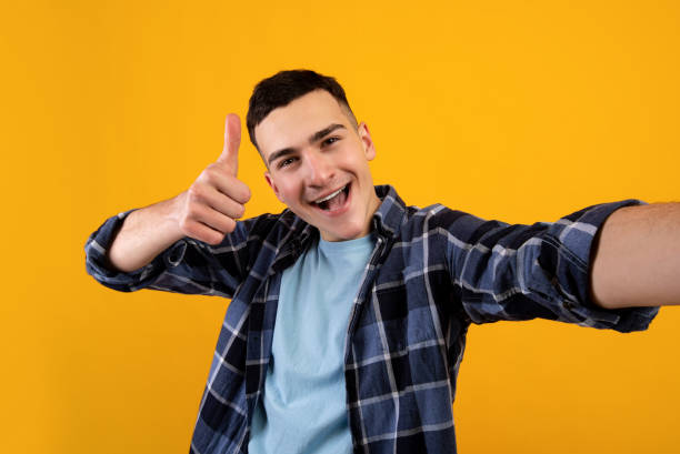istock Cool millennial guy taking selfie, showing thumb up gesture on orange studio background 1311408074