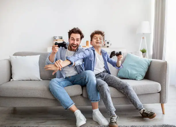 Joyful dad and son with joysticks playing video games at home, boy distracting father with hand, faving fun together, sitting on sofa in living room. Fatherhood and leisure activities concept