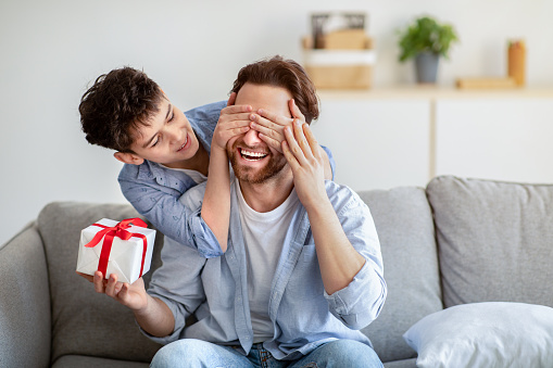 Happy father's day. Son congratulating his daddy, covering his eyes from behind, greeting and giving present at home. Family holiday celebration, surprise present for dad