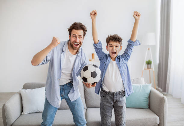 fans de football émotifs. papa et fils gais encourageant avec la bille de football, regardant le match à la tv - soccer child indoors little boys photos et images de collection