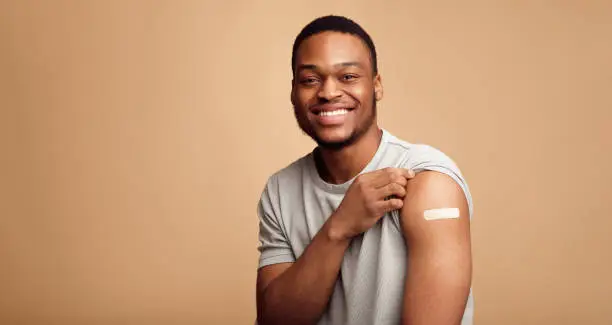Covid-19 Vaccination. Portrait Of Happy Vaccinated African Man Showing His Arm After Coronavirus Antiviral Vaccine Shot Over Beige Background. Covid Immunization Campaign Concept. Panorama