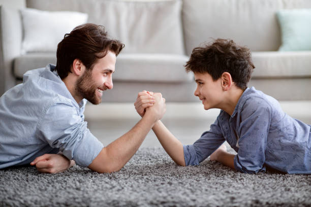 happy father competing in arm-wrestling with his son, enjoying time together at home, side view - arm wrestling imagens e fotografias de stock