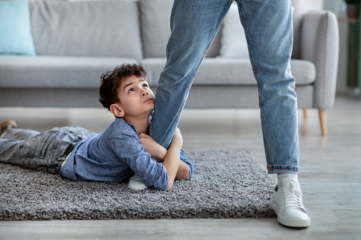 Please stay, dad. Sad little boy embracing his father leg and looking up with begging gaze while lying on the floor carpet. Upset son holding daddy before go to work