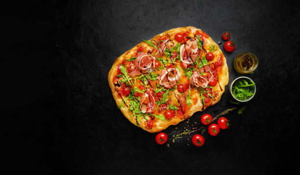 istock Roman square flatbread pizza with ham and cherry tomatoes on black slate background, top view, copy space 1311410554