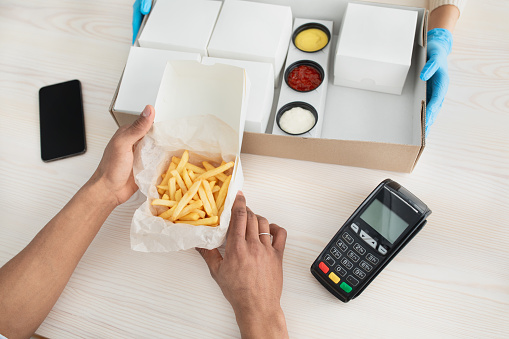 Junk food, online order and delivery during COVID-19 epidemic. Hands of courier in protective gloves give box, client looks at fries, near phone with empty screen and terminal, top view, cropped