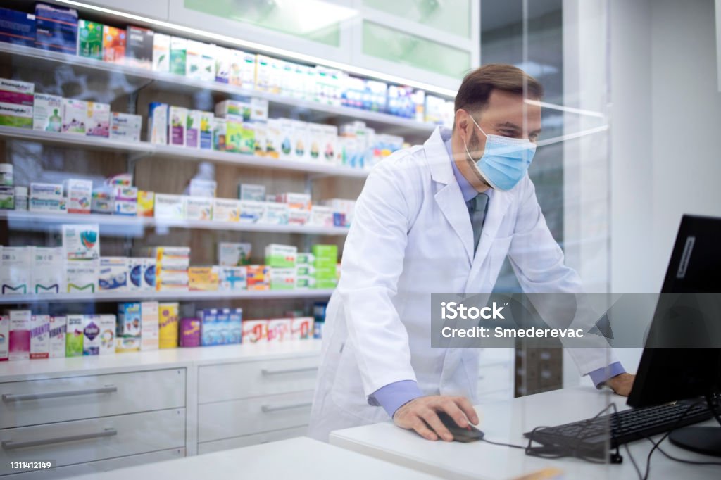 Pharmacist wearing face protection mask and white coat selling vitamins in pharmacy store during corona virus pandemic. Pharmacy Stock Photo
