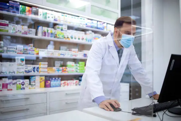 Pharmacist wearing face protection mask and white coat selling vitamins in pharmacy store during corona virus pandemic.
