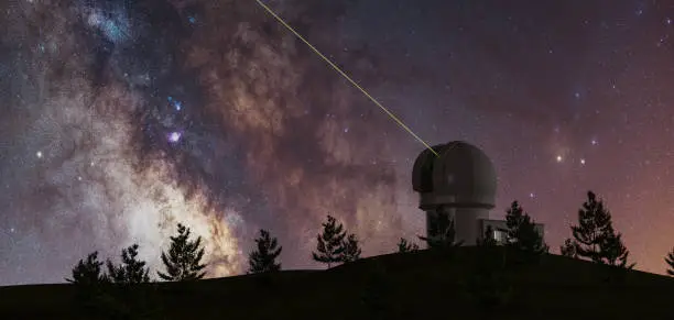 Photo of milky way with large telescope on the horizon and pine trees