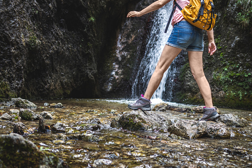 Woman with hiking boots walking on rocks in river next to waterfall. Tourist trekking at mountain trail