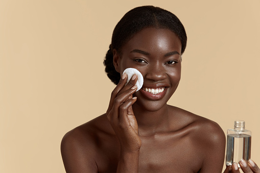 Portrait close up of beautiful black girl touch her clean face with cotton pad. Smiling young woman hold face tonic. Concept of face skin care. Isolated on beige background. Studio shoot
