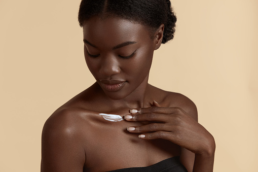 Portrait close up of beautiful black girl apply cosmetic cream on body. Serious young woman with perfect skin. Concept of skincare. Isolated on beige background. Studio shoot