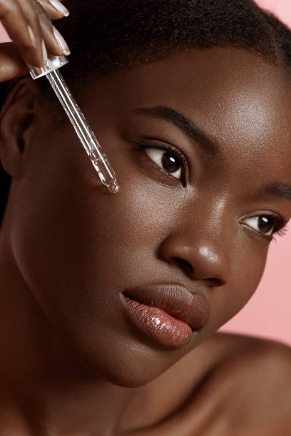 Girl dropping serum collagen moisturizer on face Portrait close up of beautiful black girl dropping serum collagen moisturizer on face. Serious young woman. Concept of face skin care. Isolated on pink background. Studio shoot melanin photos stock pictures, royalty-free photos & images