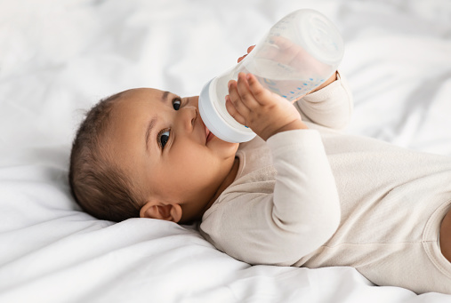 Feeding And Babycare. Closeup portrait of adorable smiling black baby holding bottle and drinking water, wearing bodysuit and resting on the white bedsheets in bedroom, lying on the back