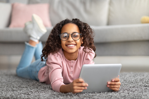 Cheerful homelanders girl wearing glasses using digital tablet, lying on carpet and smiling to camera. Teen surfing internet, studying online, checking social media, watching video or movie at home