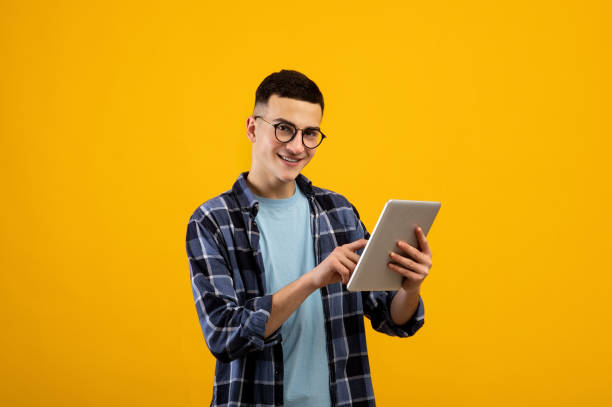 Online life concept. Funky young man using tablet pc on orange studio background Online life concept. Funky young man using tablet pc on orange studio background. Joyful millennial guy with touch pad surfing web, watching videos, checking email, posting in social media Student app stock pictures, royalty-free photos & images