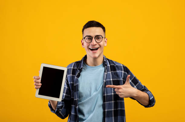 istock Cool young man pointing at blank tablet screen with space for website or app design over orange background, mockup 1311425569