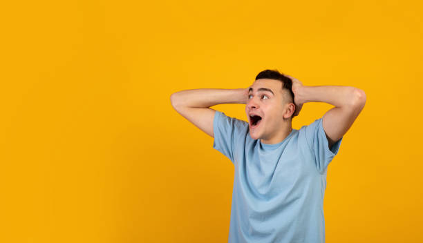 istock Cool young guy holding his head in shock, screaming OMG and looking aside at empty space on orange studio background 1311426291