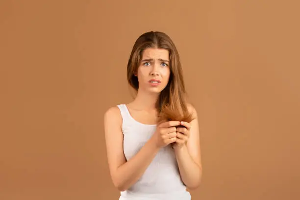 Photo of Frustrated young woman having problem with split ends on brown studio background. Beauty and wellness concept