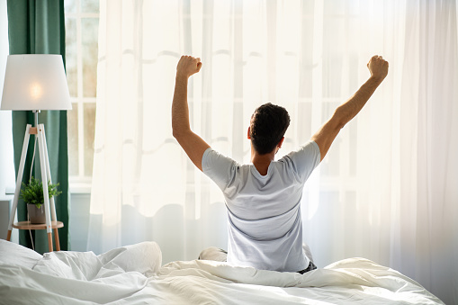 Good morning. Young man waking up and stretching his arms in cozy bedroom, back view. Guy sitting on bed facing the window, awakening after good sleep and rest