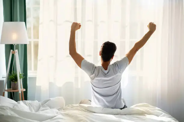 Good morning. Young man waking up and stretching his arms in cozy bedroom, back view. Guy sitting on bed facing the window, awakening after good sleep and rest