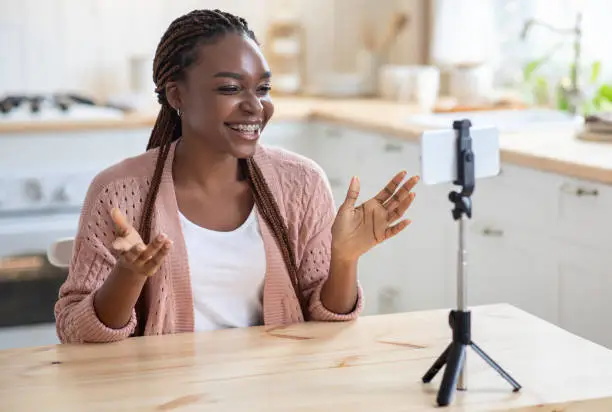 Photo of Black female recording video content for her vlog, using smartphone on tripod