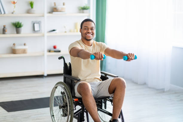 Rehabilitation of disabled people. Young African American man in wheelchair working out at home Rehabilitation of disabled people. Young African American man in wheelchair working out at home. Handicapped millennial guy training with dumbbells, doing physiotherapy exercises paraplegic stock pictures, royalty-free photos & images