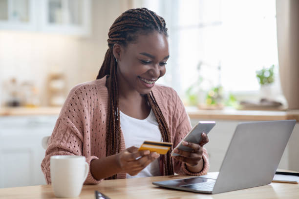 Online Payments. Cheerful black lady using smartphone and credit card in kitchen Online Payments. Cheerful black lady using smartphone and credit card in kitchen at home, smiling african woman sitting at table with laptop, paying bills in internet or transferring money via app exchanging photos stock pictures, royalty-free photos & images