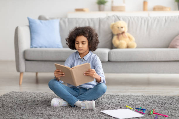istock Cute black girl sitting on floor carpet, reading book 1311430389