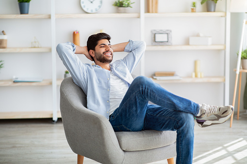 Lazy day. Relaxed arab man resting on comfortable armchair, holding hands behind head. Guy sitting with closed eyes and enjoying weekend in home interior