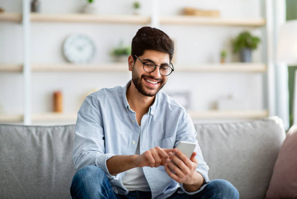 istock Checking new app. Happy arab man using smartphone, relaxing on comfy sofa at home interior 1311431425