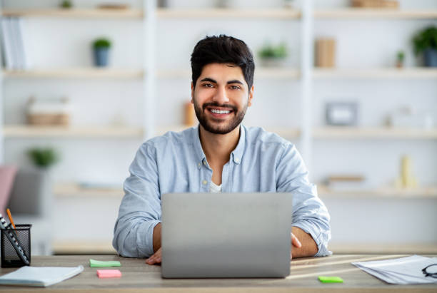 istock Positive arab male employee smiling at camera while working on laptop at home office 1311431669