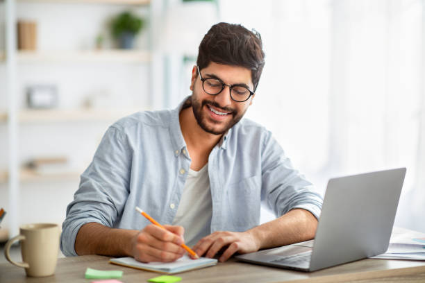 istock Happy arab man writing business report on laptop and taking notes, sitting at workplace at home interior 1311431990