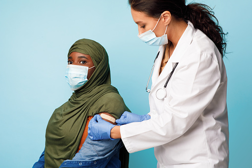 Vaccination Immunization Campaign. Doctor Applying Plaster On Vaccinated Female Patient's Arm After Making Vaccine Injection To African Muslim Lady On Blue Background. Studio Shot