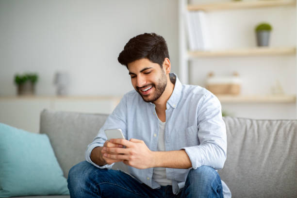 istock Cheerful arab guy sitting with smartphone on comfy sofa at home, messaging with friends 1311432254