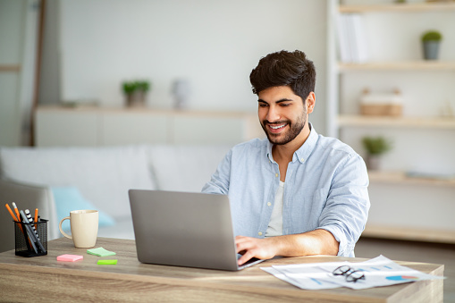 Positive freelancer working on laptop, typing on keyboard and looking at screen, checking online project. Arab man sitting at desk at home interior and enjoying remote work, copy space