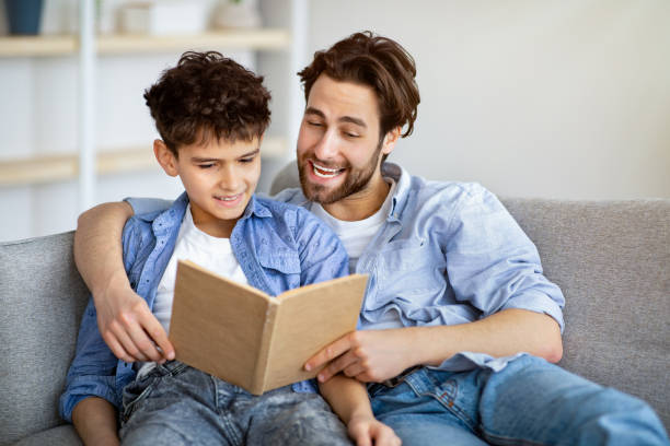 istock Useful time at home. Father and son reading book and smiling, resting together on comfy sofa. 1311433289