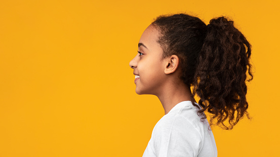 Side view profile portrait of cute African American girl with beaming smile and curly hair in ponytail looking aside at empty free copy space isolated on yellow studio background, banner, panorama
